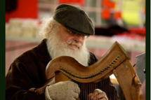 Man with white hair and beard wearing a cap and gloves plays a harp